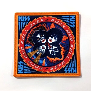 May include: A square orange fabric patch with a circular design featuring the band KISS. The design includes the band's name, "Rock and Roll Over", and four stylized faces of the band members. The background is blue with orange flames.
