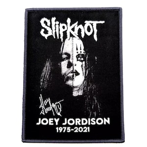 Könnte beinhalten: Ein schwarzer rechteckiger Aufnäher mit dem Bandnamen "Slipknot" in Weiß, über einem Schwarz-Weiß-Porträt von Joey Jordison. Der Aufnäher enthält seine Unterschrift und die Jahre 1975-2021.