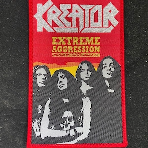 May include: A red, black, and yellow embroidered patch with the band name "Kreator" and the text "Extreme Aggression". The patch features a black and white image of four people.