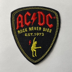 May include: Black, shield-shaped patch with yellow stitching. The red text reads "AC/DC" with a lightning bolt, "ROCK NEVER DIES" and "EST. 1973". A yellow figure is playing guitar. A classic rock and roll emblem.