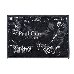 Może przedstawiać: Czarna i biała prostokątna naszywka z wizerunkiem muzyka grającego na gitarze basowej. Naszywka zawiera tekst "Paul Gray 1972-2010" oraz nazwę zespołu "Slipknot" w stylizowanej czcionce, a także podpis.