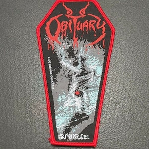 Puede incluir: Parche bordado rojo y negro con forma de ataúd con el nombre de la banda "Obituary" en letras rojas y el texto "Live In Bang Cock 2013" en letras blancas.