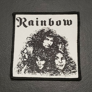 Op de afbeelding: Een witte stoffen patch met zwarte geborduurde tekst die "Rainbow" luidt en een zwart-witte afbeelding van een band.