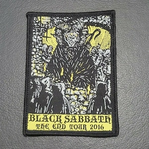 May include: Black Sabbath The End Tour 2016 embroidered patch. The patch features a black and white illustration of a figure with horns and a staff, surrounded by other figures. The background is a yellow-gold colour.