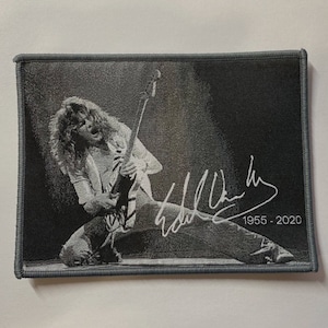 Peut inclure: Un écusson brodé en noir et blanc représentant un musicien jouant de la guitare avec une signature et les dates 1955 - 2020.