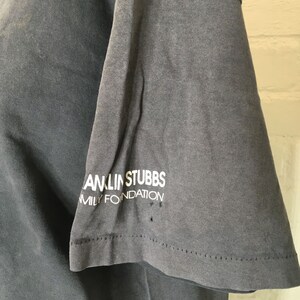 Puede incluir: Primer plano de una manga de camiseta gris con el texto blanco "ANLIN STUBBS FAMILY FOUNDATION". La tela muestra signos de desgaste, con peque&ntilde;os agujeros cerca del dobladillo. La camiseta parece vintage.