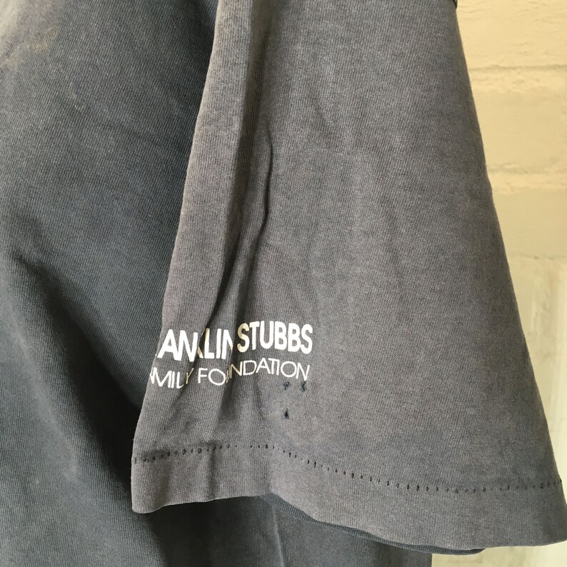 Puede incluir: Primer plano de una manga de camiseta gris con el texto blanco "ANLIN STUBBS FAMILY FOUNDATION". La tela muestra signos de desgaste, con peque&ntilde;os agujeros cerca del dobladillo. La camiseta parece vintage.