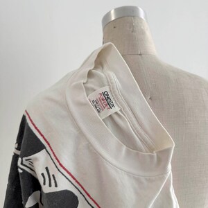 May include: White t-shirt with a black and red graphic design. The shirt is a crew neck and appears to be a vintage item. The tag reads "ONEITA POWER-T". The shirt is displayed on a mannequin.