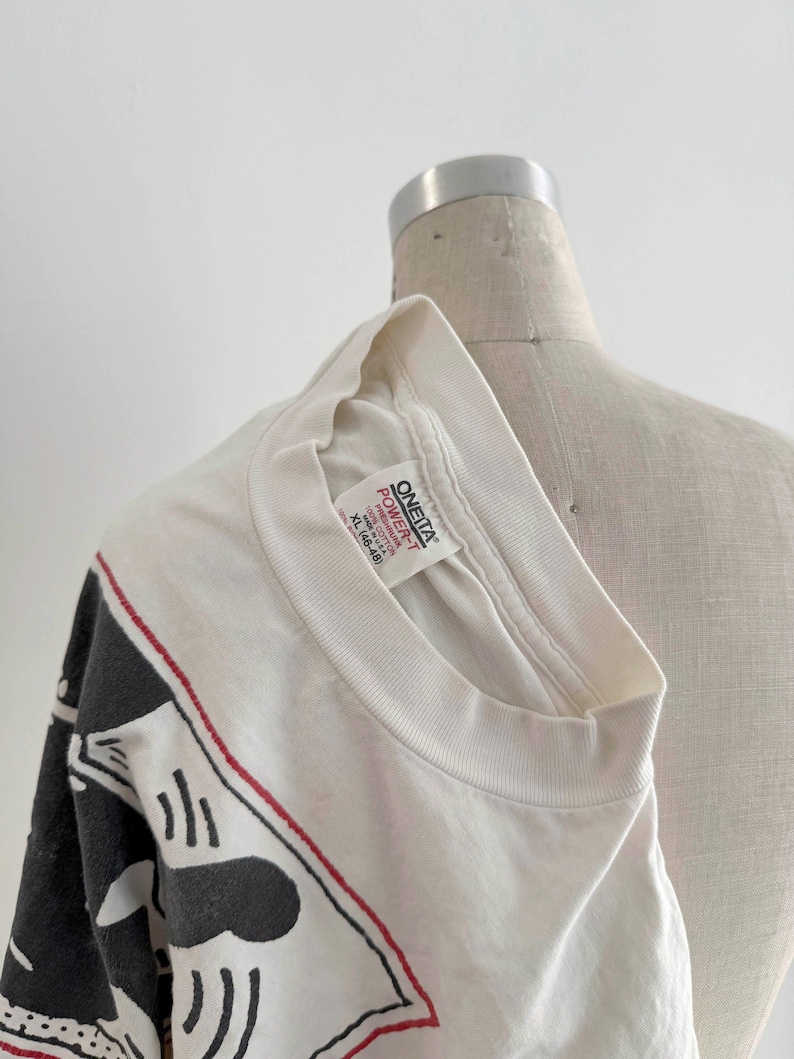 May include: White t-shirt with a black and red graphic design. The shirt is a crew neck and appears to be a vintage item. The tag reads "ONEITA POWER-T". The shirt is displayed on a mannequin.