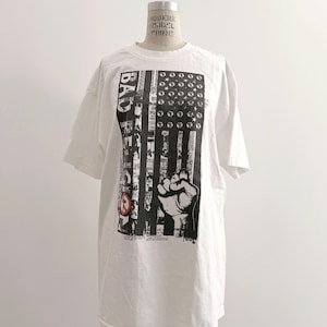 May include: White t-shirt with a graphic design featuring a black and white American flag with a clenched fist. The word "BAD" is printed vertically on the left side of the flag. The shirt is displayed on a mannequin.