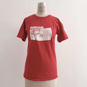 May include: A red t-shirt with short sleeves, displayed on a mannequin. The shirt features the text "SNAPCASE" in white block letters above a white graphic with a barcode design. The shirt is made of a soft material.