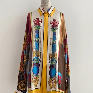 Op de afbeelding: Een lang mouw overhemd met knopen en een kleurrijk, gepatroneerd ontwerp. Het overhemd heeft een gele streep in het midden en verschillende afbeeldingen, waaronder engelen, kolommen en mythische wezens. Het overhemd is gemaakt van een zijdeachtige stof.