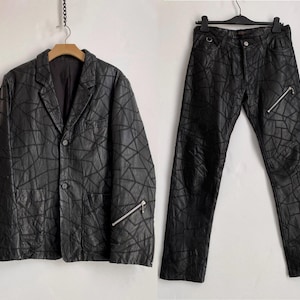 May include: Black blazer and trousers set. The blazer features a two-button fastening, notched lapels, and two front pockets. The trousers have a zip closure and a distinctive geometric pattern. Both items have silver zips.