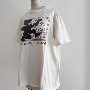 May include: Off-white t-shirt featuring a black and red graphic design with the text "FREE SOUTH AFRICA". The graphic depicts a stylized figure and other shapes within a rectangular frame. The shirt has short sleeves and a crew neck.