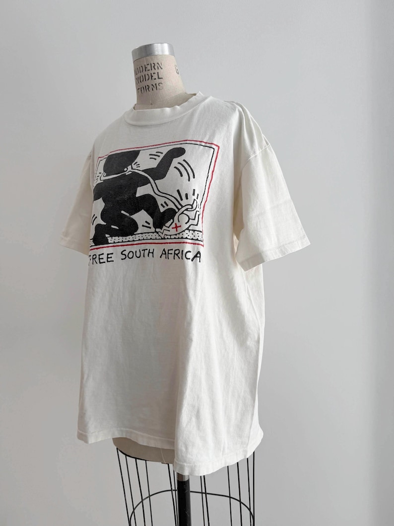 May include: Off-white t-shirt featuring a black and red graphic design with the text "FREE SOUTH AFRICA". The graphic depicts a stylized figure and other shapes within a rectangular frame. The shirt has short sleeves and a crew neck.