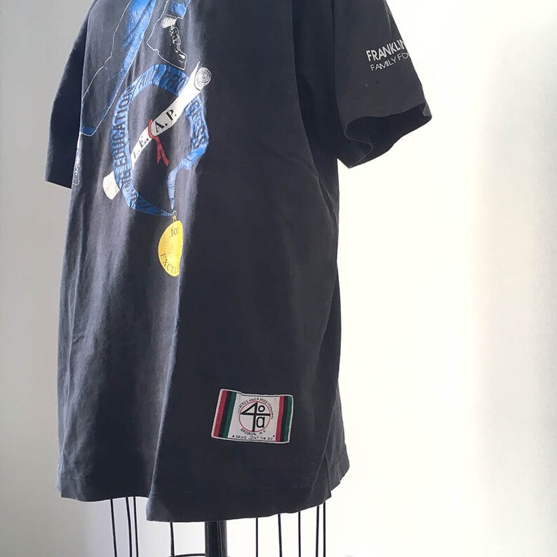 Puede incluir: Camiseta negra con un gr&aacute;fico en la espalda que presenta un dise&ntilde;o azul y blanco con texto. Un parche con texto y un dise&ntilde;o de rayas rojas, verdes y blancas est&aacute; en la parte inferior derecha. La camiseta se muestra en un maniqu&iacute;.