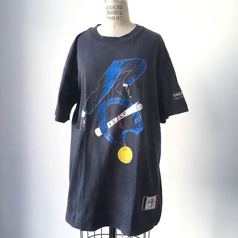 Puede incluir: Camiseta negra con un gr&aacute;fico de una persona corriendo con pantalones azules y un diploma. La camiseta tiene el texto "I.E.A.P." y una medalla de oro. La camiseta tambi&eacute;n tiene el texto "Franklin Family Fund".