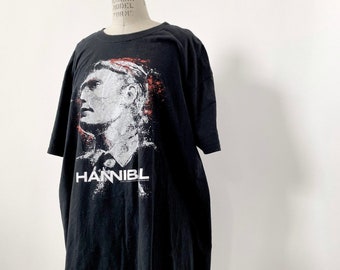 May include: Black t-shirt featuring a white and red graphic of a man's face and the word "HANNIBAL" printed on the front.