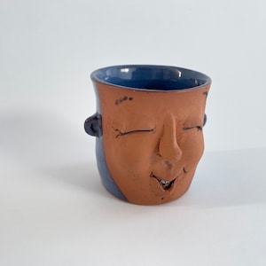 May include: A ceramic mug with a face sculpted on the front. The mug is glazed in a blue color on the top half and a brown color on the bottom half. The face has closed eyes and a smiling mouth.