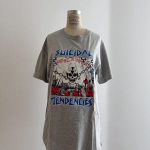 May include: A heather gray short-sleeve t-shirt featuring a graphic print of a horned skull with chains, red hearts, and the text Suicidal Tendencies and Wont Fall In Love Today. The shirt is displayed on a mannequin torso with a neutral background.