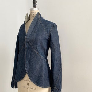 Pode incluir: Casaco de ganga azul escuro com um design único, com gola alta e fecho de um botão. O casaco é exibido num manequim, mostrando o seu ajuste sob medida e os intrincados detalhes das costuras. O casaco tem uma bainha curva e mangas compridas.