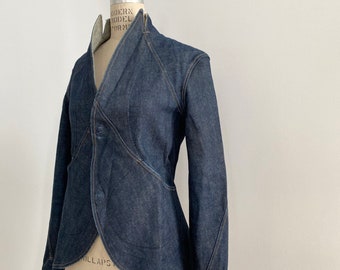 May include: A dark blue denim jacket with a unique design, featuring a high collar and a single-button closure. The jacket is displayed on a mannequin, showcasing its tailored fit and intricate seam details. The jacket's design includes a curved hemline and long sleeves.