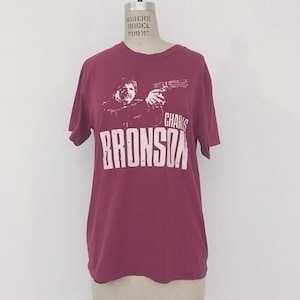 Puede incluir: Camiseta burdeos con un gráfico blanco de un hombre sosteniendo un arma, con el nombre "CHARLES BRONSON" impreso debajo. La camiseta se muestra en un maniquí. Tiene mangas cortas y cuello redondo.