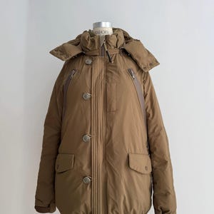 May include: A brown winter coat with a hood, button fastenings, and multiple pockets. The coat is displayed on a mannequin. The coat has a quilted design and a high collar. The coat has two zipped pockets on the chest.
