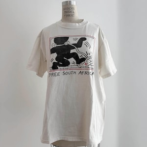 May include: Off-white t-shirt featuring a black graphic of a figure with a line drawing style, with the words "FREE SOUTH AFRICA" in black. The graphic is framed with a red border. The shirt is displayed on a mannequin.