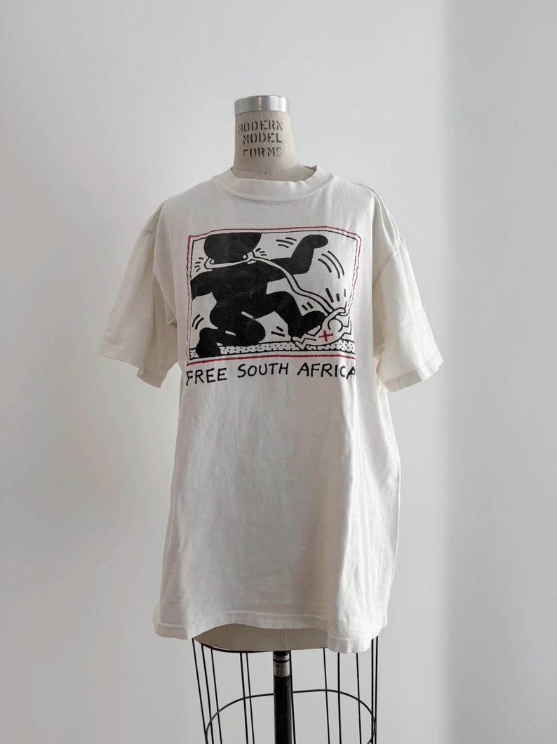 May include: Off-white t-shirt featuring a black graphic of a figure with a line drawing style, with the words "FREE SOUTH AFRICA" in black. The graphic is framed with a red border. The shirt is displayed on a mannequin.