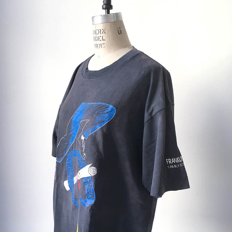 Puede incluir: Camiseta gris oscuro con un gr&aacute;fico azul y blanco de una persona sosteniendo un pergamino. La manga tiene el texto "FRANKLIN FAMILY FUN". La camiseta se muestra en un maniqu&iacute;.