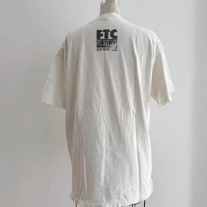 May include: Cream-colored t-shirt with short sleeves, displayed on a mannequin. The back of the shirt features black text that reads "FTC PRO SKATE SHOP SAN FRANCISCO". The shirt has a relaxed fit and a classic crew neck.