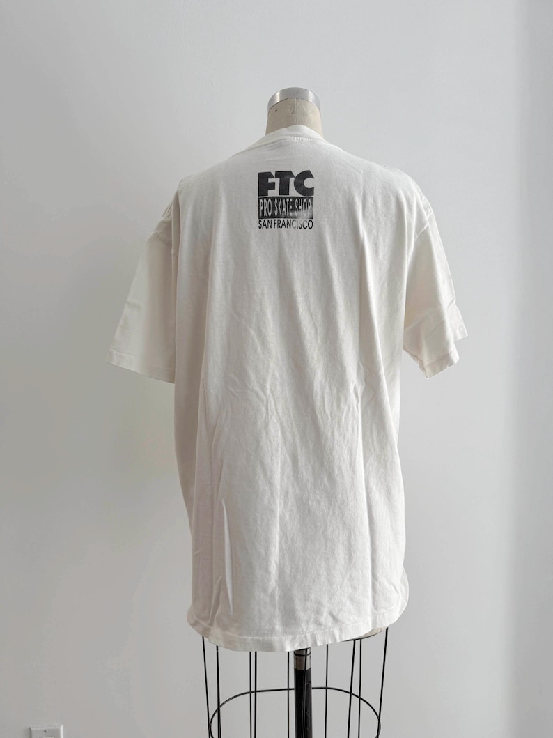 May include: Cream-colored t-shirt with short sleeves, displayed on a mannequin. The back of the shirt features black text that reads "FTC PRO SKATE SHOP SAN FRANCISCO". The shirt has a relaxed fit and a classic crew neck.