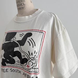 May include: Off-white t-shirt featuring a black graphic of a figure with a red border and the text "FREE SOUTH AFRICA". The short-sleeved shirt is displayed on a mannequin.