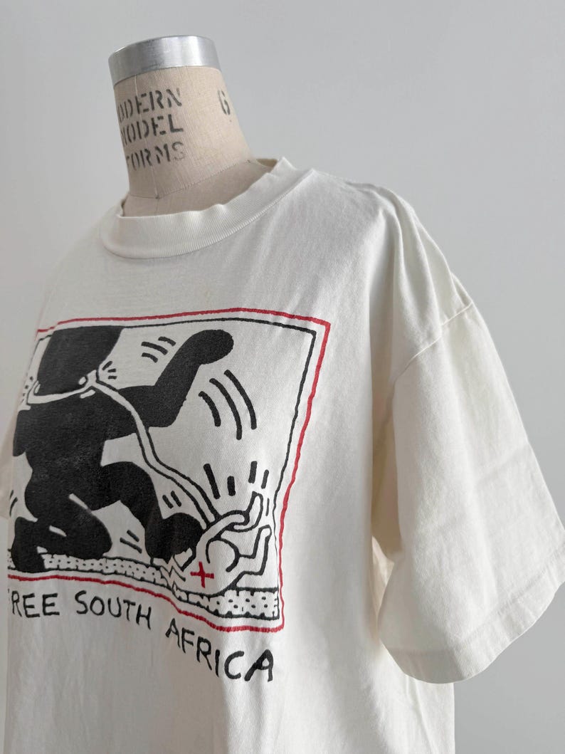 May include: Off-white t-shirt featuring a black graphic of a figure with a red border and the text "FREE SOUTH AFRICA". The short-sleeved shirt is displayed on a mannequin.