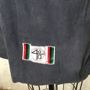 Puede incluir: Camiseta gris oscuro con un parche rectangular en la esquina inferior derecha. El parche presenta el texto "40" dentro de un c&iacute;rculo, con texto adicional y rayas rojas, verdes y negras.