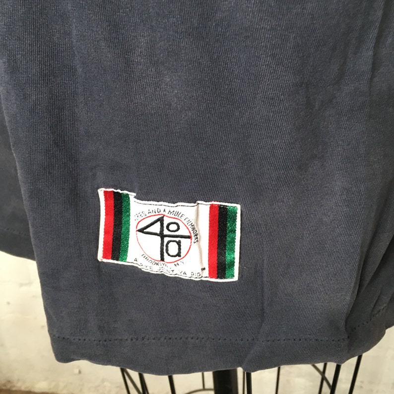 Puede incluir: Camiseta gris oscuro con un parche rectangular en la esquina inferior derecha. El parche presenta el texto "40" dentro de un c&iacute;rculo, con texto adicional y rayas rojas, verdes y negras.