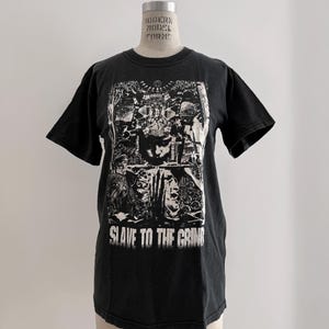 May include: Black t-shirt with a distressed graphic print and the text "SLAVE TO THE GRIND" in white. The graphic features multiple faces and figures. The shirt is displayed on a mannequin.
