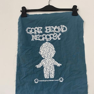 May include: A teal blue fabric patch with white text that reads "GORE BEYOND NECROPSY" and a white silhouette of a person made up of the same text. The text is repeated multiple times to form the silhouette. The bottom of the patch has the text "Arnaldrillingrind Harshit Core!!!" in white.