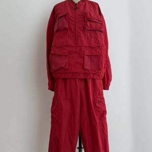May include: A red two-piece outfit on a mannequin. The top is a long-sleeved jacket with multiple front pockets and a zip. The matching trousers have cargo pockets and a zip closure. The outfit is made of a lightweight material.