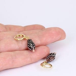 May include: Gold-colored hoop earrings with a small, black and white spotted seashell charm. The earrings are displayed against a light background. The seashell charms have a natural, organic shape.