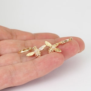 May include: A pair of gold-toned hoop earrings featuring small, detailed bee charms. Each bee charm has textured wings and a striped body, suspended from a thin, circular hoop. The metal has a polished finish, reflecting light across the surface.