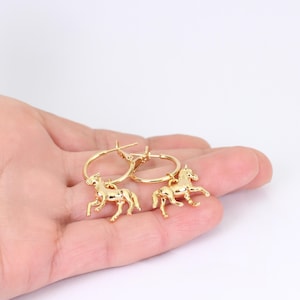 May include: A pair of gold-colored hoop earrings. Each earring features a small, detailed horse charm. The earrings are displayed against a light background, highlighting their shiny, metallic finish. The horse charms are in a running pose.