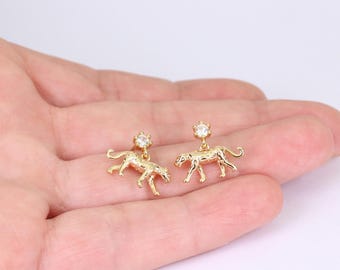 May include: A pair of gold-colored earrings featuring a small leopard design. Each earring has a clear, round gemstone at the top. The earrings are displayed on a light background.