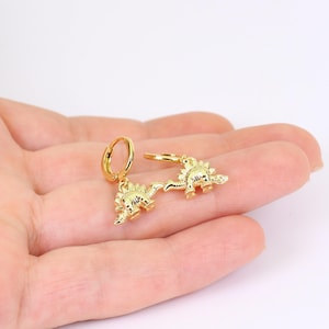 May include: A pair of gold-toned hoop earrings featuring dangling dinosaur charms. Each charm is shaped like a stegosaurus with a textured surface and a polished finish. The hoops are circular and connect to the dinosaur pendants at the top.