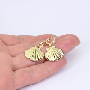 Könnte beinhalten: Ein Paar goldfarbene Muschelohrringe. Jeder Ohrring hat ein detailliertes, fächerförmiges Design mit einem Klappbügelverschluss. Die Ohrringe werden vor einem neutralen Hintergrund präsentiert.