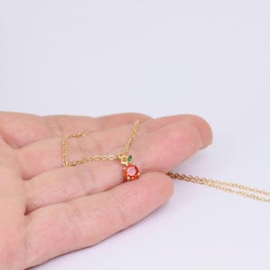 May include: A gold necklace with an apple-shaped pendant. The pendant features a round, red gemstone and small green leaves. The necklace is displayed against a white backdrop. The pendant is approximately 1 cm in diameter.