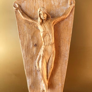 以下が含まれることがあります： 腕を広げた人物の彫刻が施された木製の壁飾り。写実的なスタイルで彫られています。彫刻は薄い茶色の仕上げの板に取り付けられています。