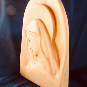 以下が含まれることがあります： ドレープされた頭部覆いを持つ穏やかな女性像を描いた、彫刻された木製のレリーフ彫刻。彫刻は明るい木の色調で、人物の横顔とハローのようなアーチがレリーフで彫られています。背景は濃い青色です。