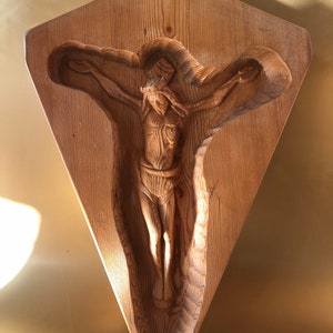 May include: A wooden wall sculpture of a carved figure of Jesus Christ on the cross. The sculpture is in a diamond shape and is made of light brown wood.